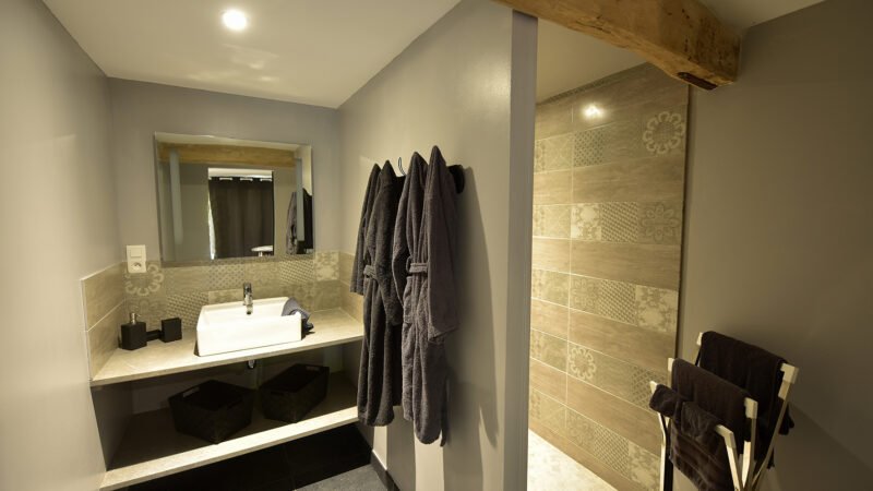 Salle de bain élégante et moderne du gîte Le Clos de la Rose, avec douche italienne, peignoirs, vasque contemporaine et poutre apparente.