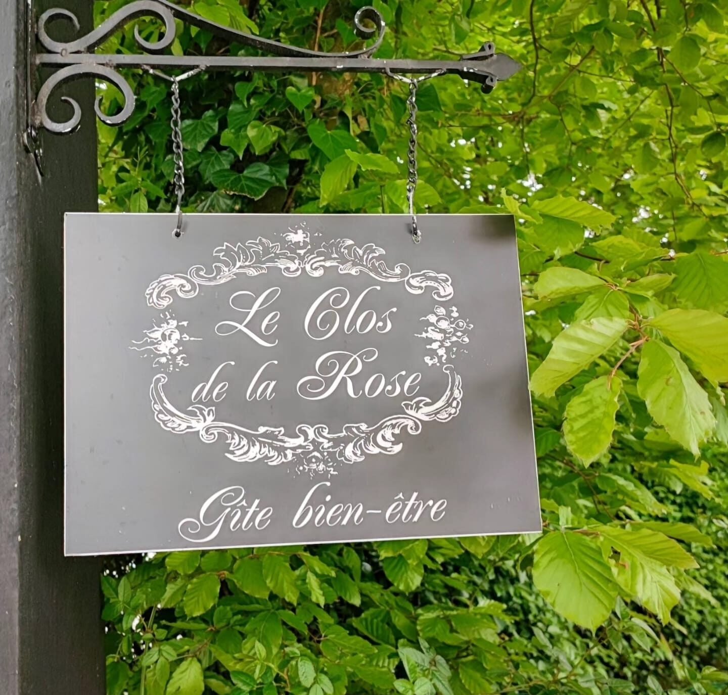 Panneau en fer forgé du gîte Le Clos de la Rose, suspendu à un support décoratif, indiquant un hébergement bien-être dans un cadre verdoyant.